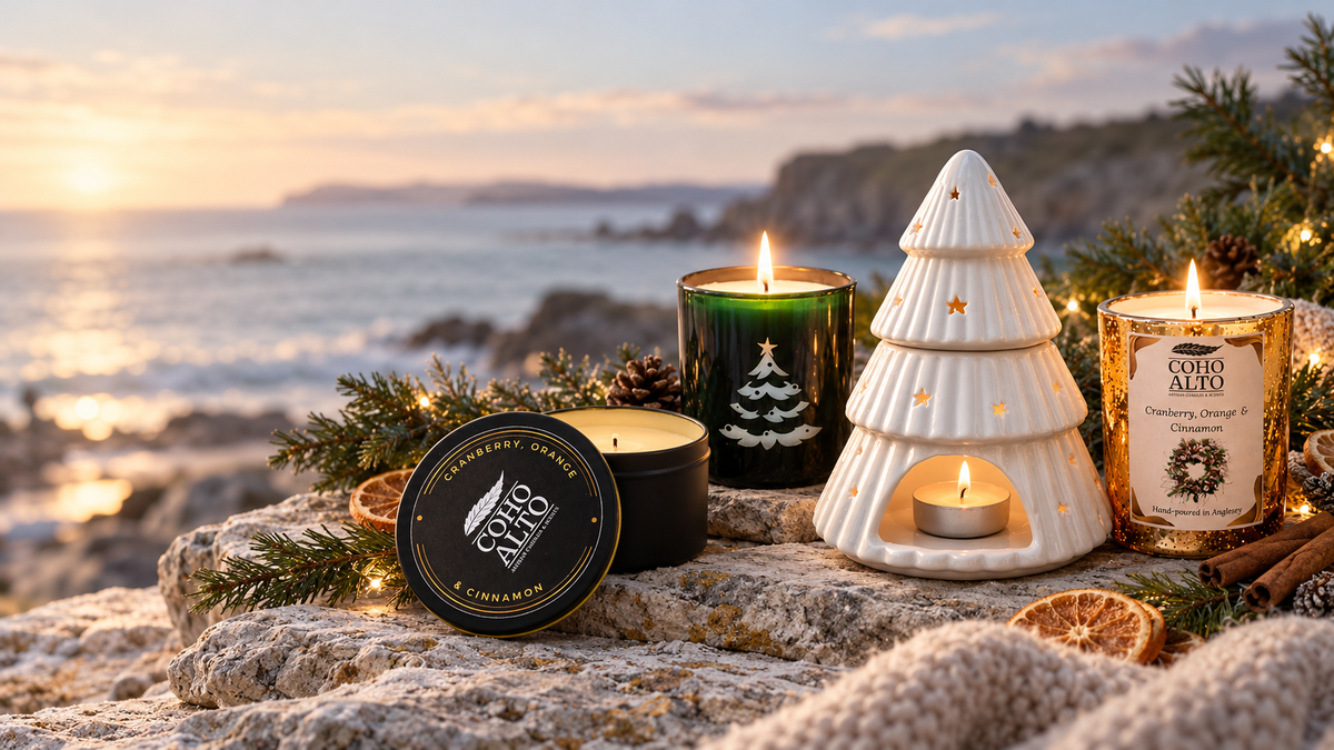 A set of Coho Alto holiday candles arranged on a rocky cliffside overlooking the ocean at sunset. The set includes a black tin candle labeled 'Cinnamon', a green candle with a white Christmas tree design, a white ceramic Christmas tree-shaped holder with a tea light inside, and a gold candle labeled 'Cinnamon Spice'.